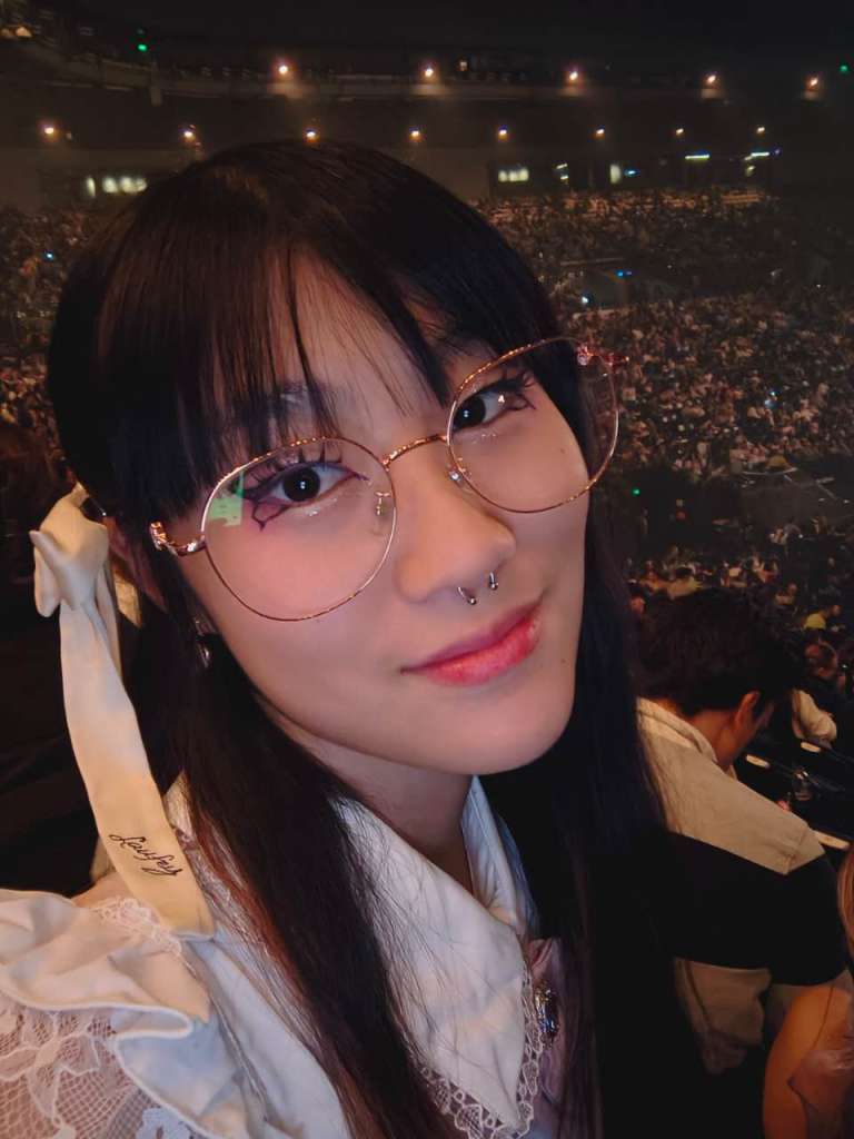 23-year-old woman with black hair, a fringe and glasses sitting in stadium. 